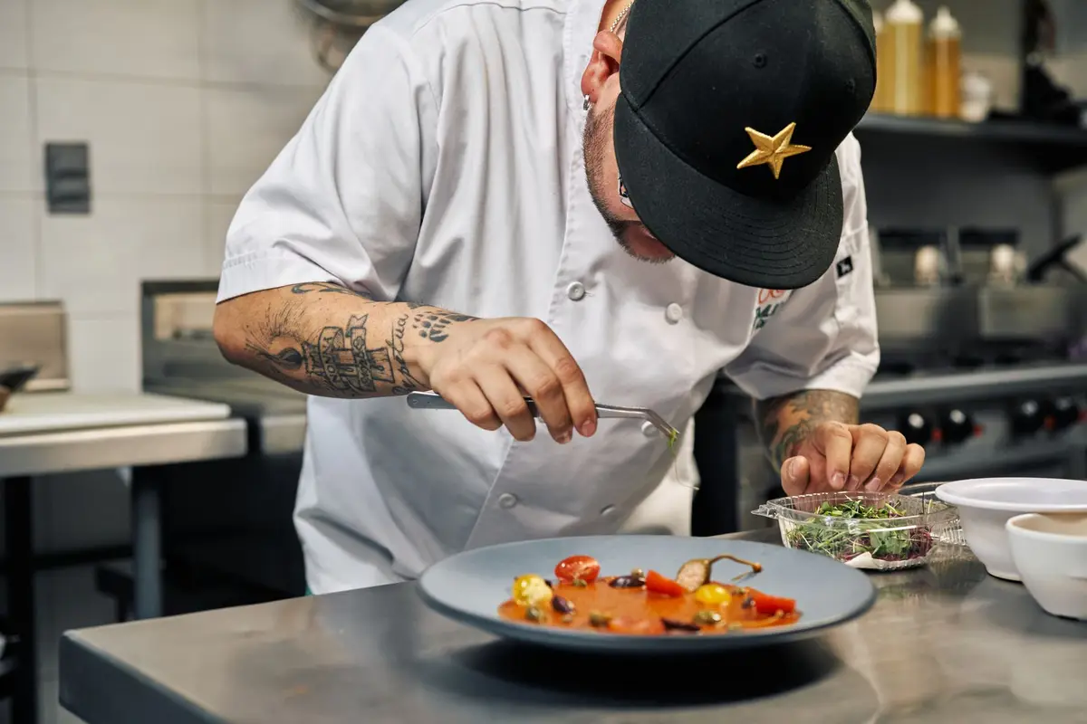 Tattooed chef in cap and whites plating a dish with tweezers at a restaurant kitchen pass