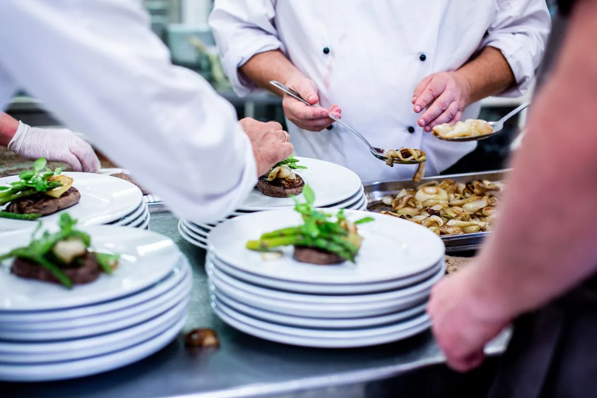 Chefs in whites plating a course onto stacks of white plates on a steel kitchen pass